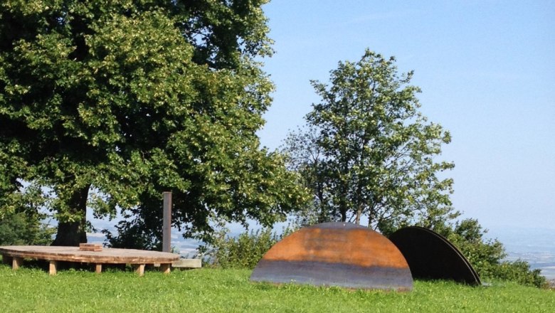 Holzplattform und Metallskulpturen auf einer Wiese mit Bäumen im Hintergrund.