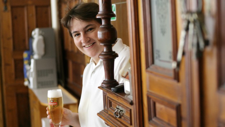 Person in einem Gasthaus mit einem Glas Bier in der Hand, lächelnd hinter einer Holzbar.
