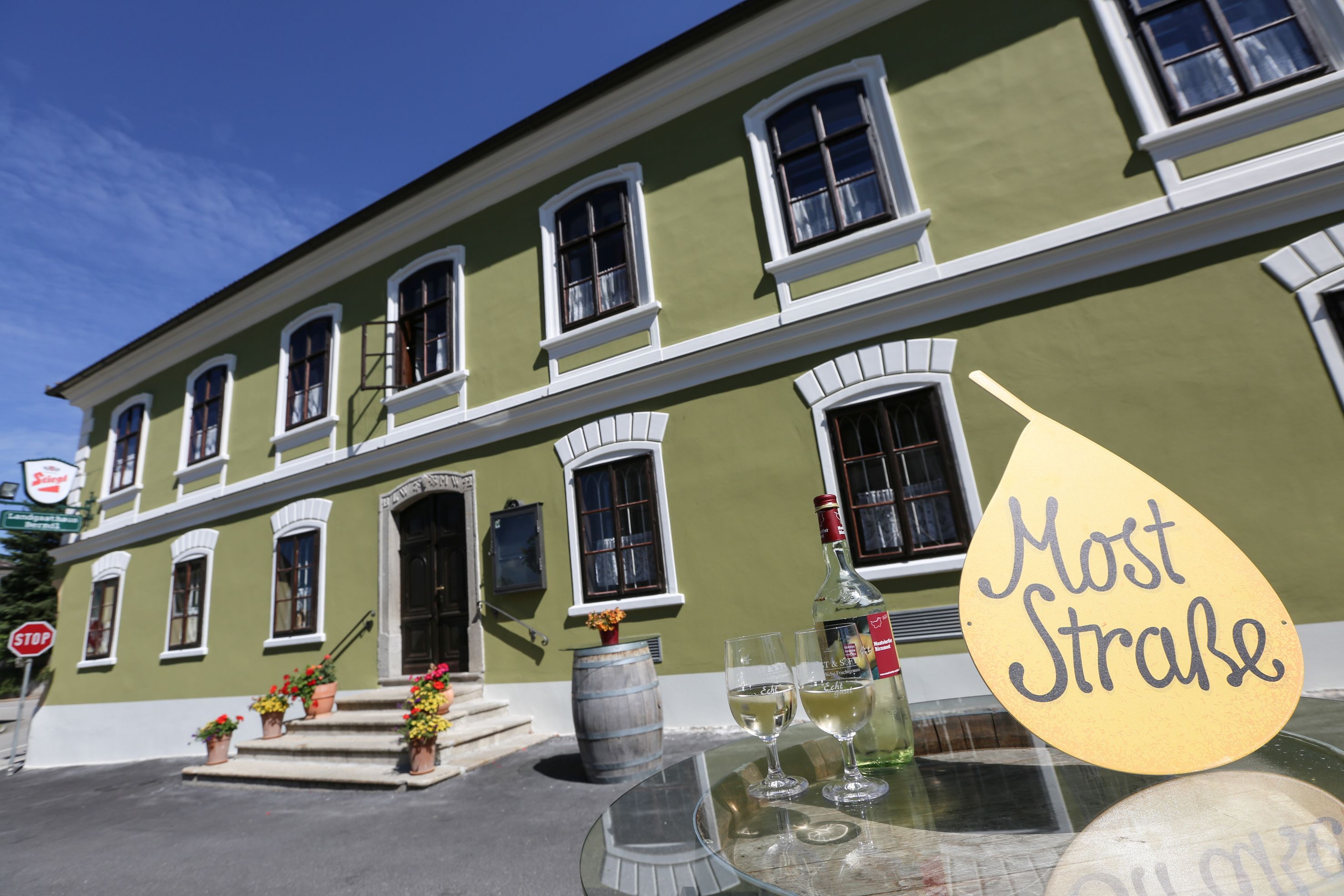 Grünes Gebäude mit weißen Fenstern, davor ein Tisch mit Weinflasche und Gläsern, Schild mit Aufschrift 'Most Straße'.
