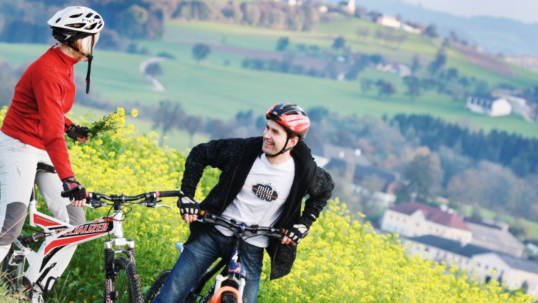 Zwei Radfahrer mit Helmen stehen auf einem blühenden Feld mit grüner Landschaft im Hintergrund.