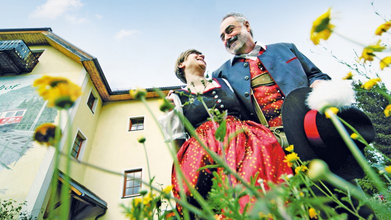 Ein Paar in traditioneller Tracht steht vor einem gelben Gebäude, umgeben von Blumen, und lächelt sich an.