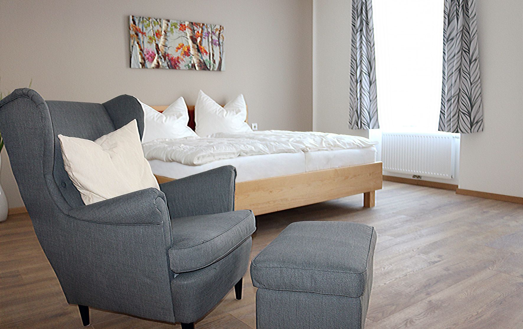 Grauer, gemütlicher Stuhl im Vordergrund. Im Hintergrund ein Doppelbett mit weißer Bettwäsche.