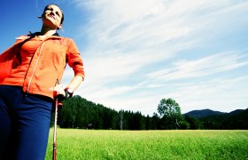 Frau beim Nordic Walking auf einer Wiese mit Wald und Bergen im Hintergrund.