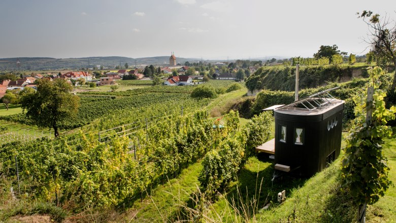 A small, modern caravan stands in the middle of vineyards with a view of a village and hills in the background.