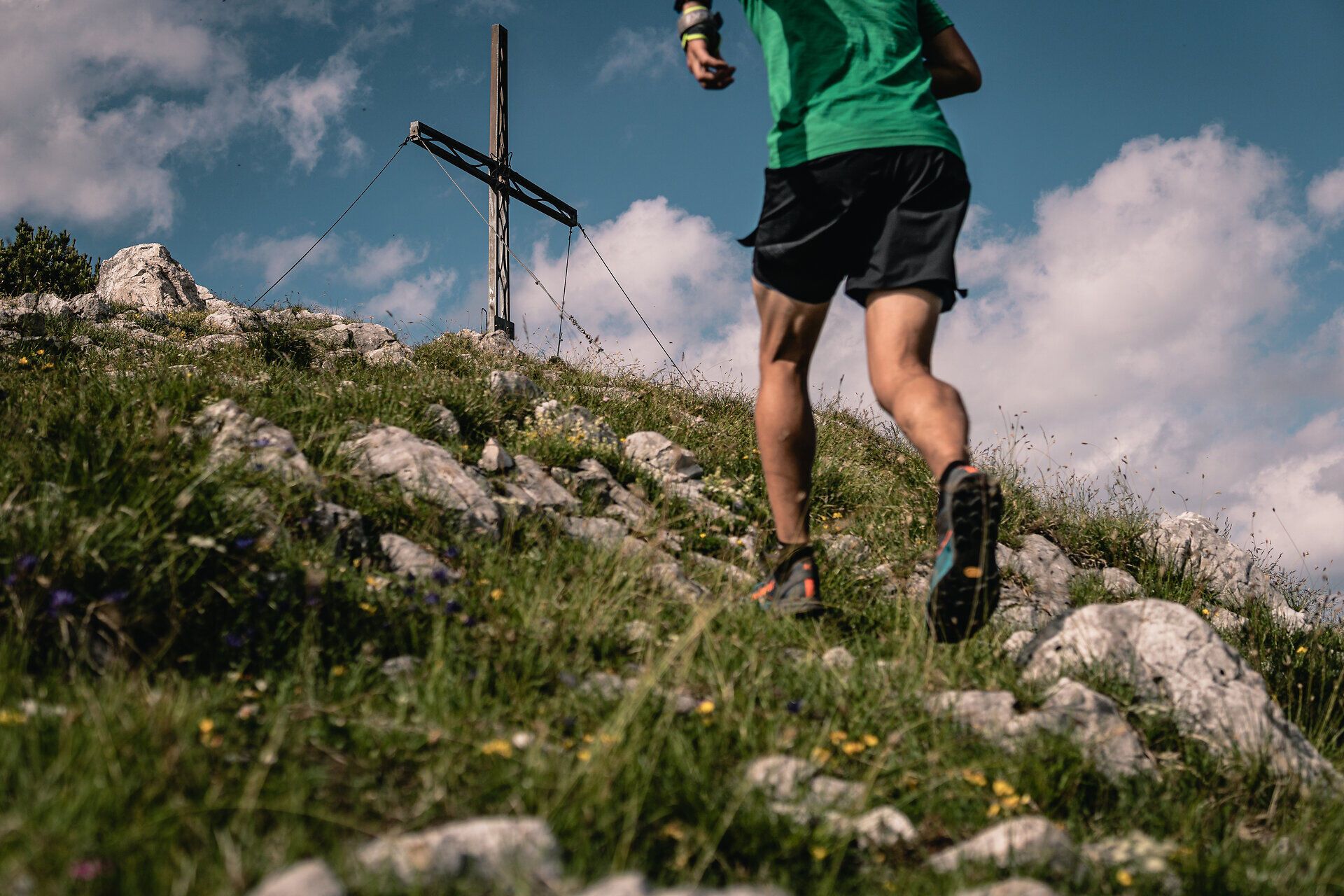 Ein Läufer erklimmt den steilen Pfad, umgeben von üppigem Grün und bunten Wildblumen. Das Gipfelkreuz kündigt den Höhepunkt des Abenteuers an, während die frische Bergluft die Sinne belebt. Hier, in der majestätischen Kulisse der Alpen, wird der Sommer in seiner schönsten Form erlebbar.