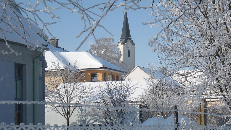 Verschneite Dorfansicht mit Kirche und Bäumen im Winter.