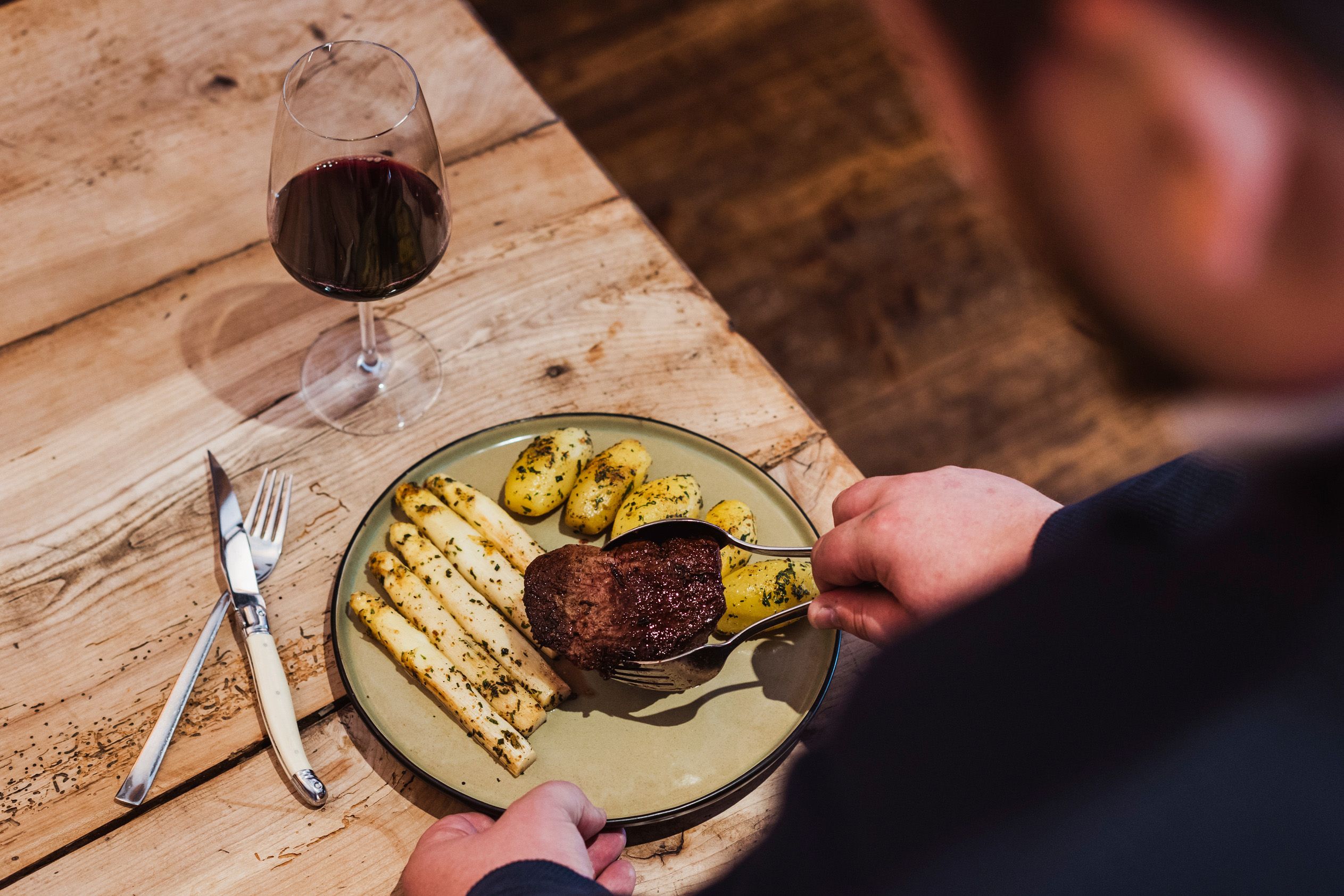 Ein Teller mit Rinderfilet, Spargel und Kartoffeln, daneben ein Glas Rotwein auf einem Holztisch.
