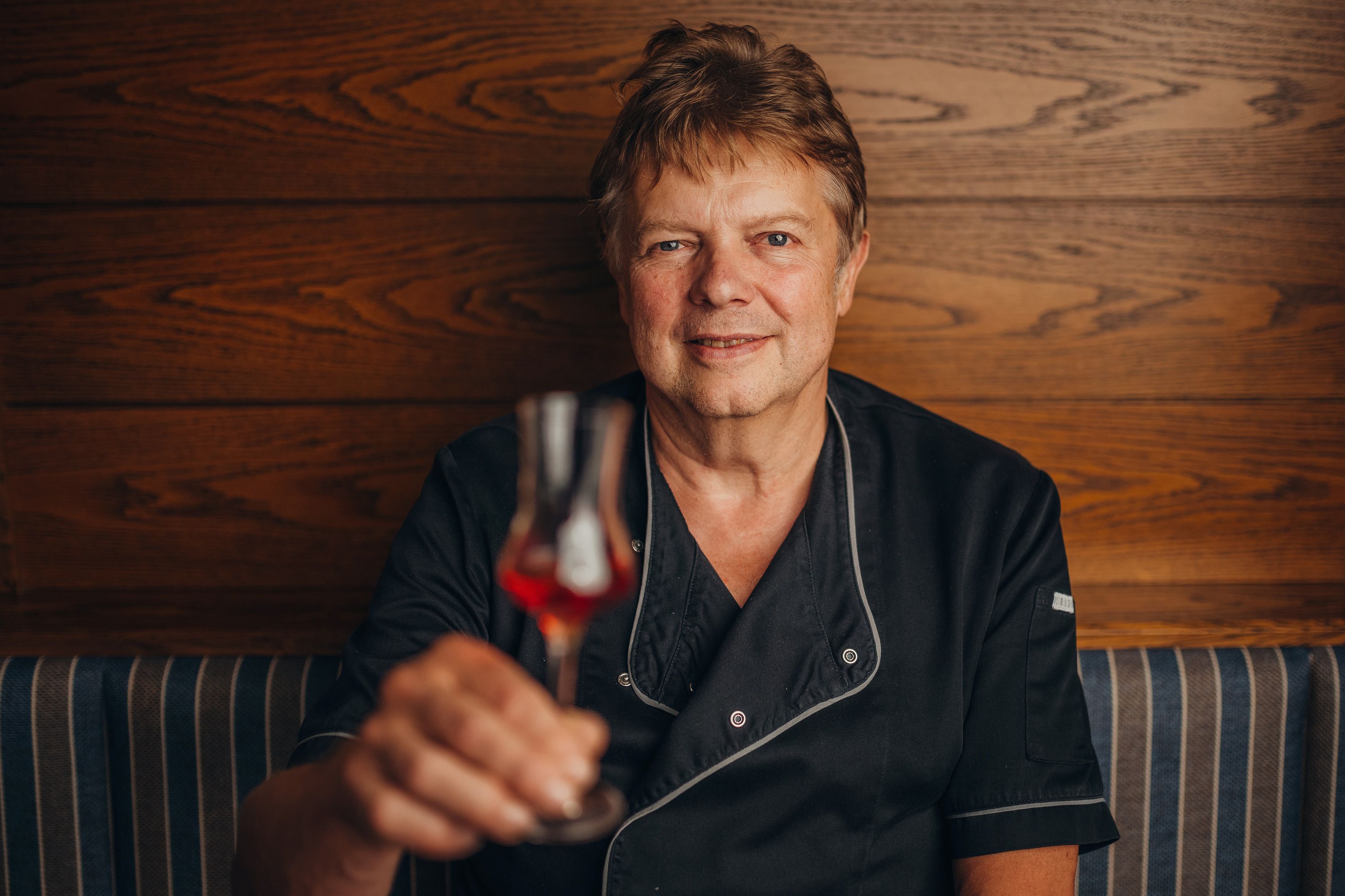 Ein Mann in schwarzer Kleidung hält ein Glas mit rotem Getränk vor einer Holzwand.