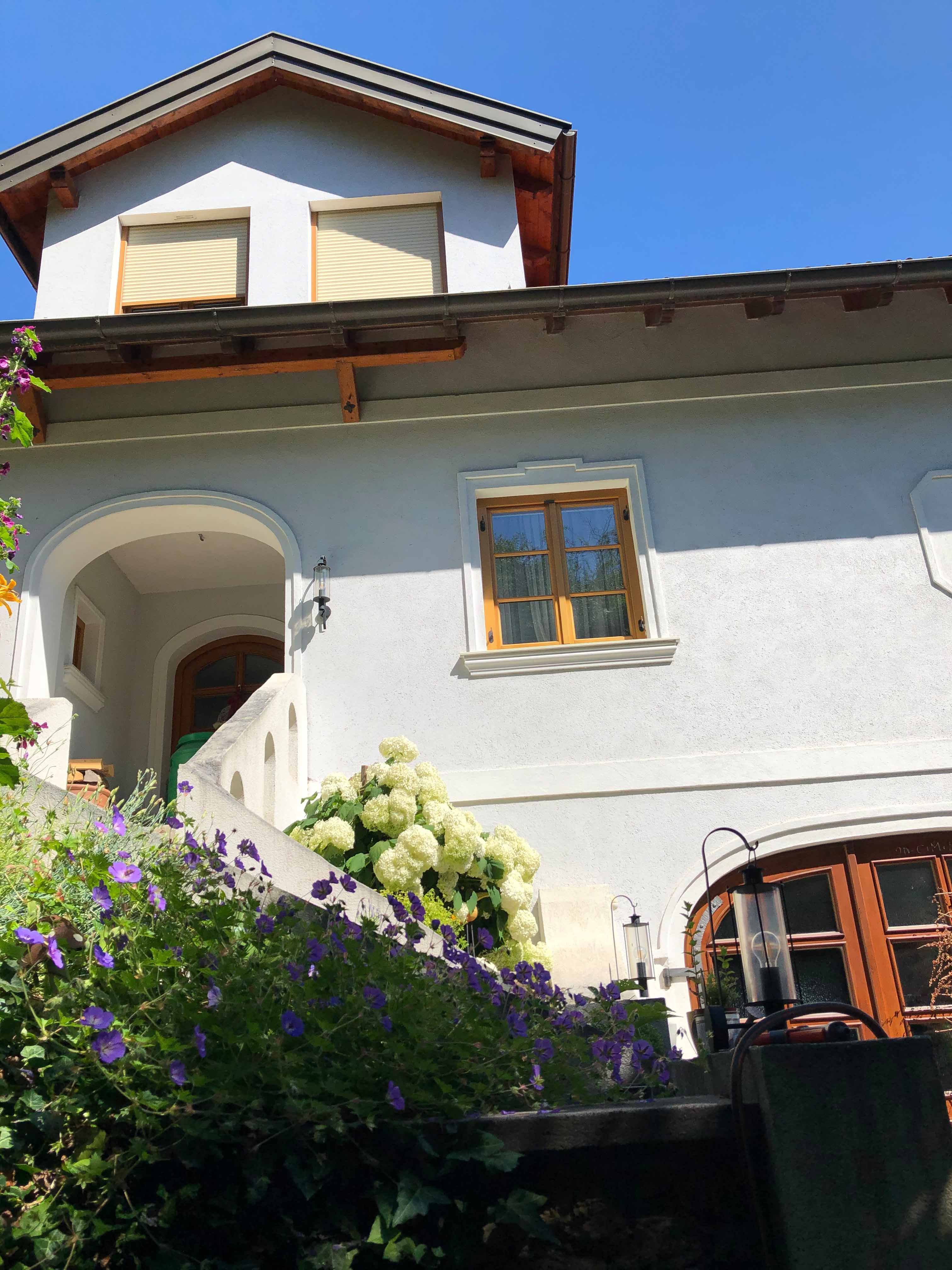 Haus mit weißen Wänden, Holzdach und Blumen im Vordergrund.