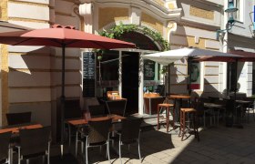 Au&szlig;enbereich eines Restaurants mit Tischen, St&uuml;hlen und Sonnenschirmen in einer sonnigen Gasse.