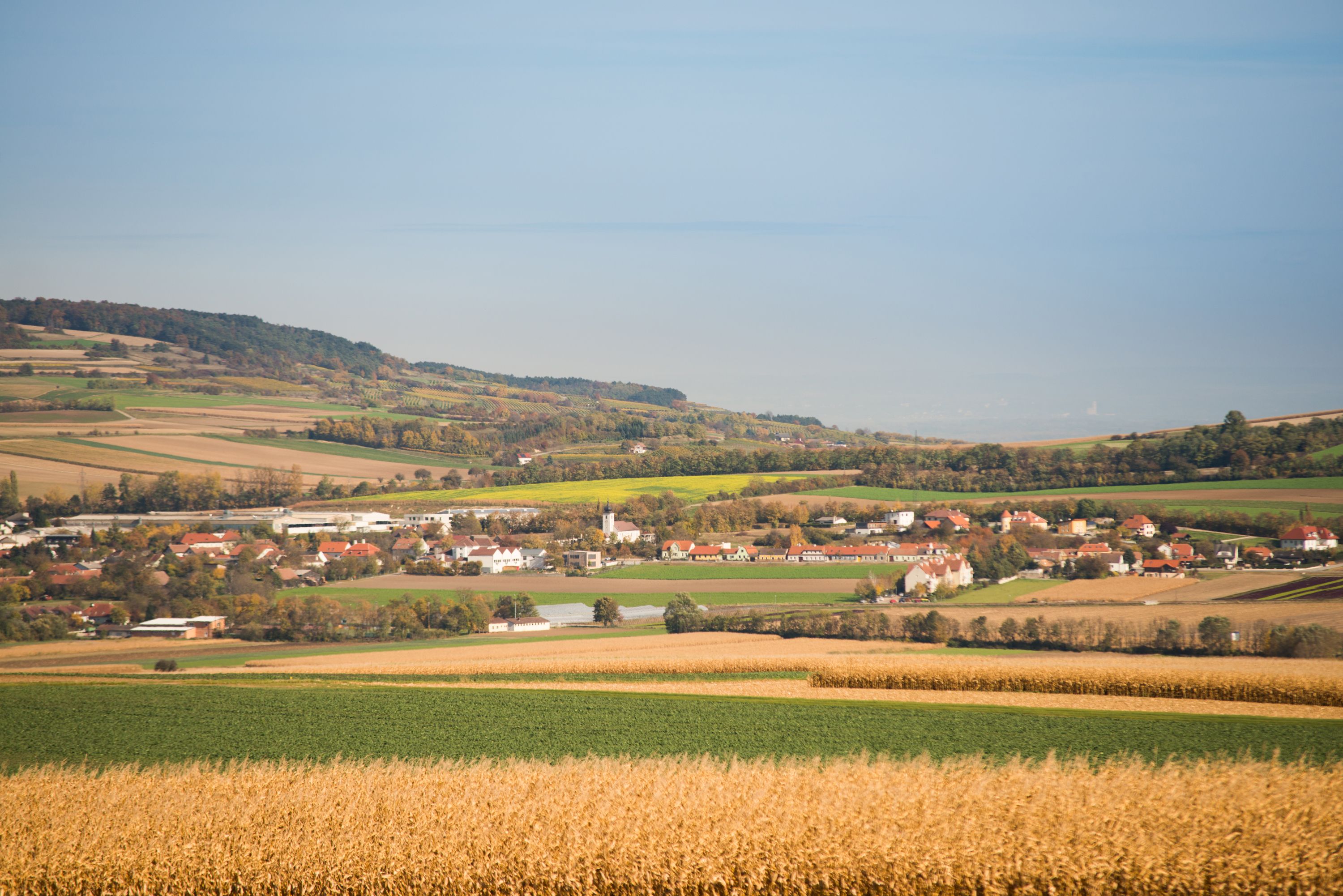 Landschaft mit Feldern und einem Dorf im Hintergrund, umgeben von Hügeln und Bäumen.