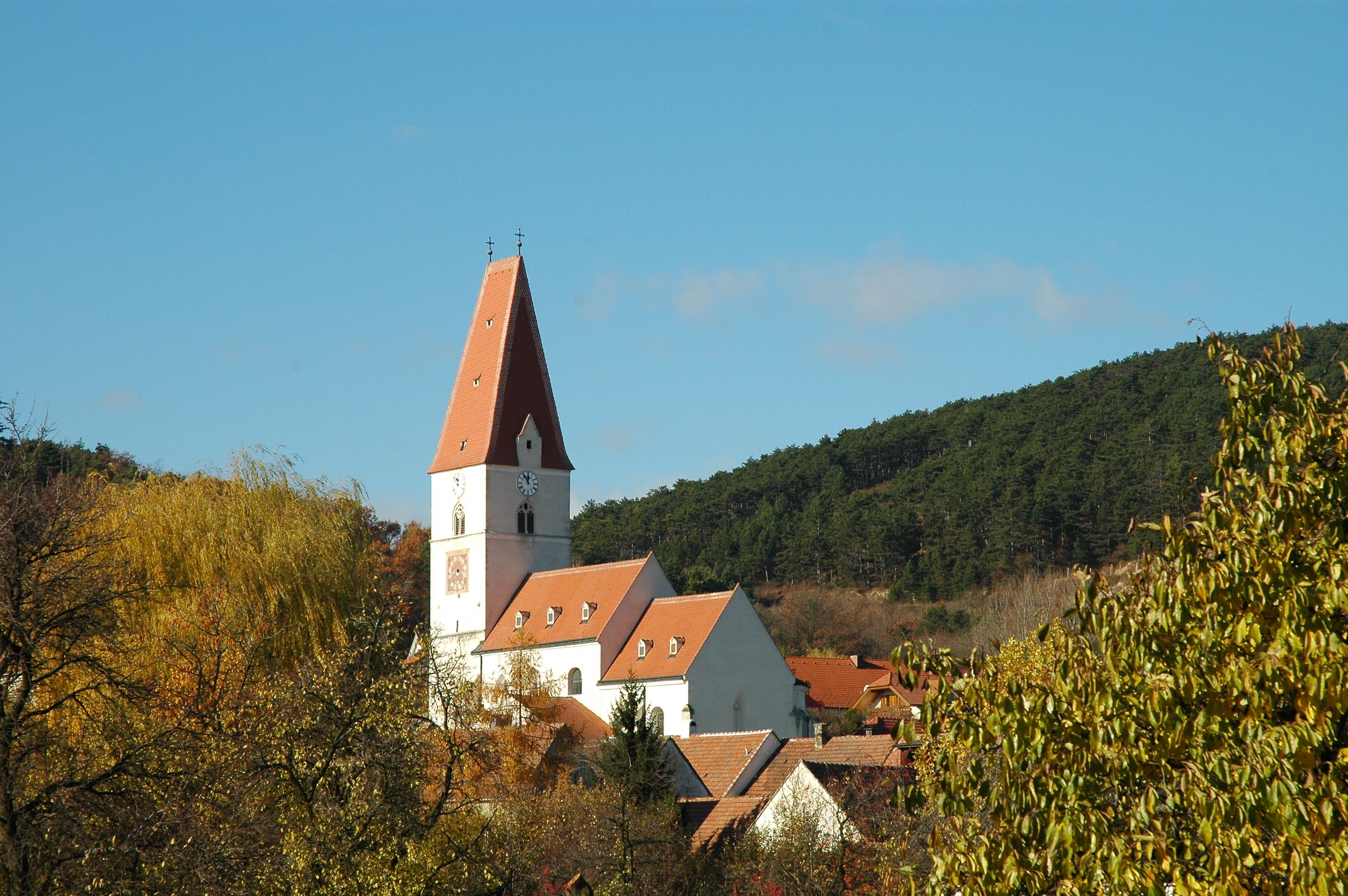 Pfarrkirche Nußdorf mit rotem Dach und Uhrturm, umgeben von Bäumen und Hügeln.