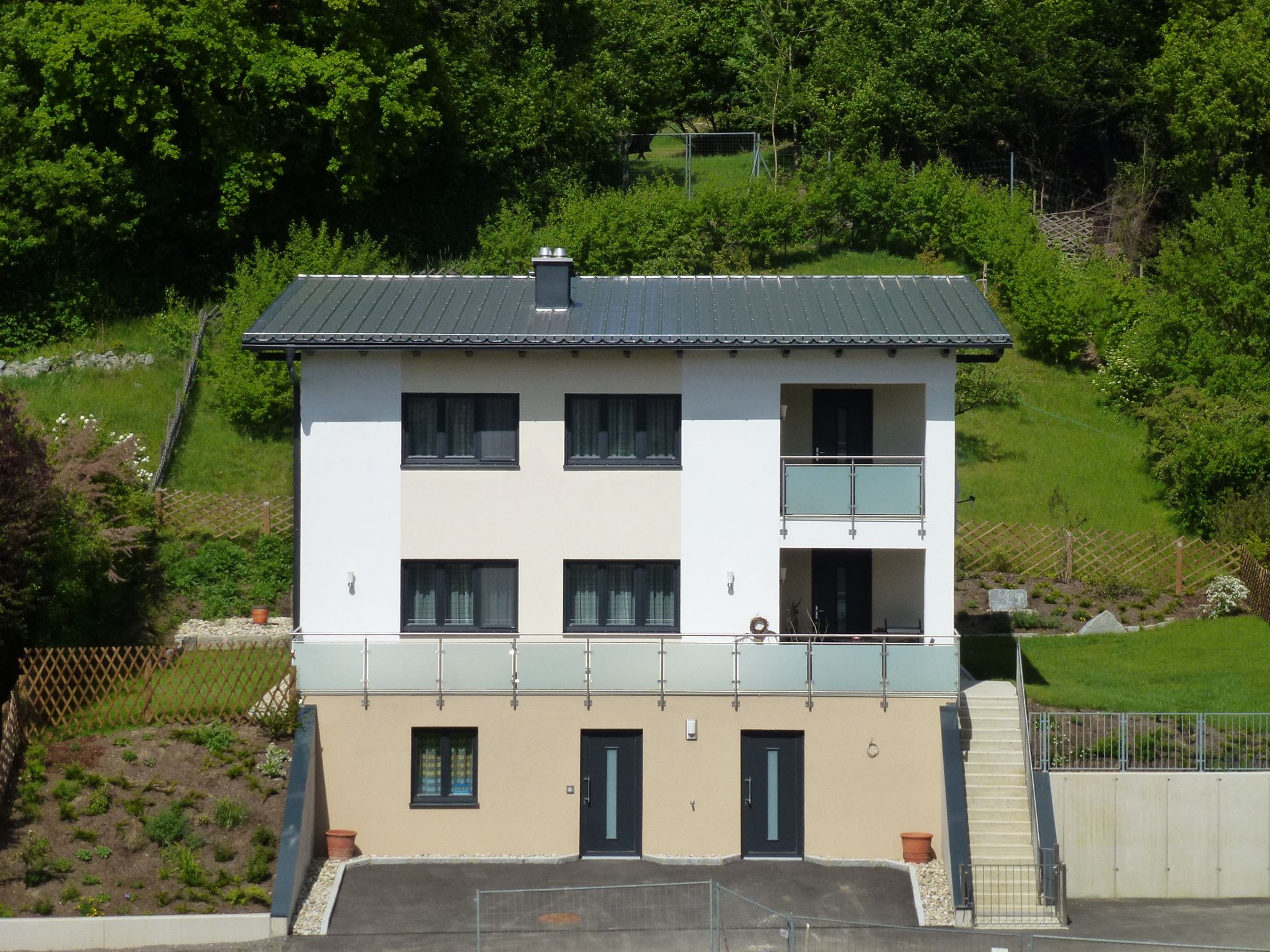 Zweistöckiges modernes Haus mit Balkon, umgeben von grüner Landschaft.