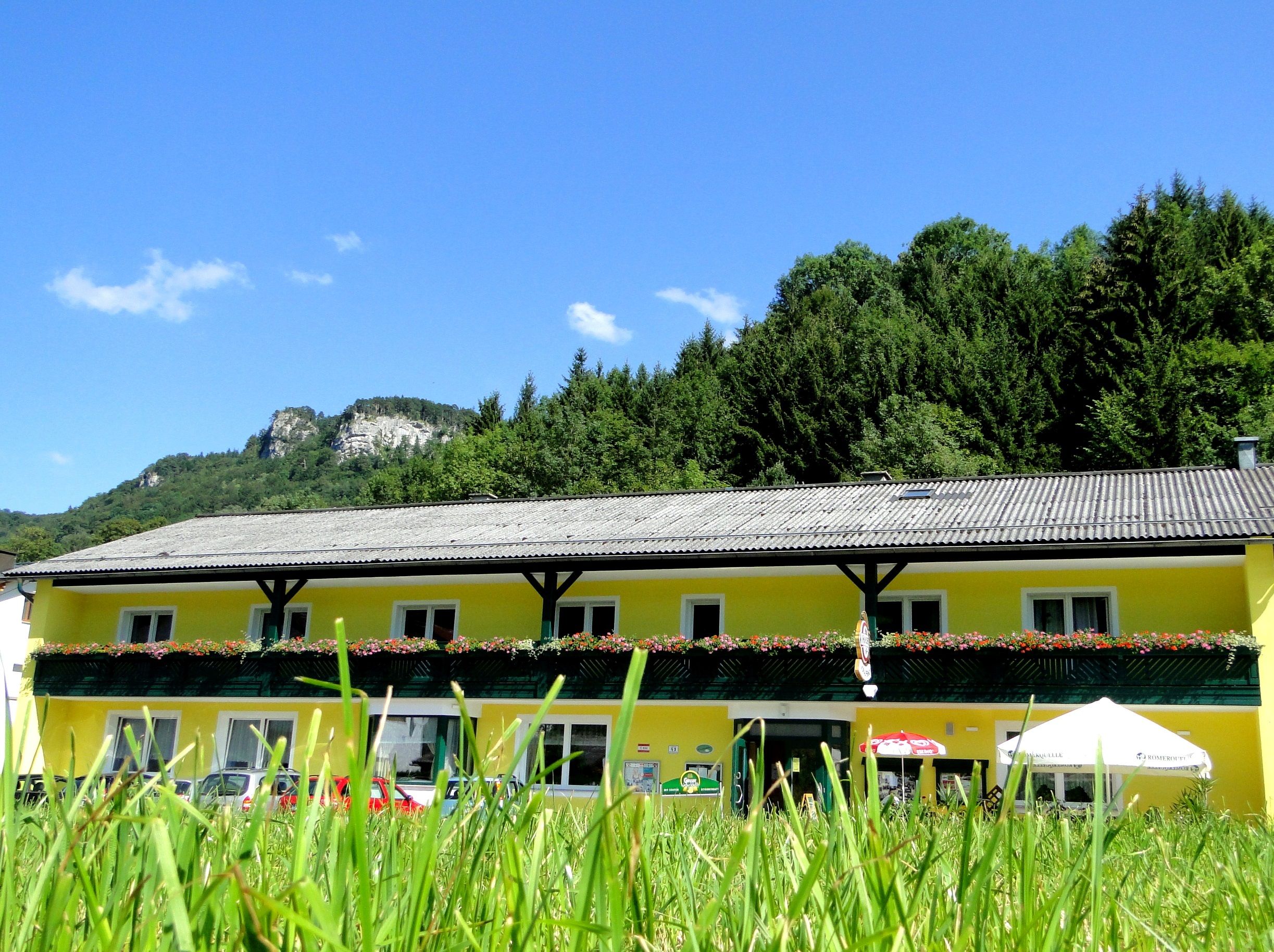 Gelbes Gebäude mit Blumenbalkon vor bewaldetem Hügel und blauem Himmel.