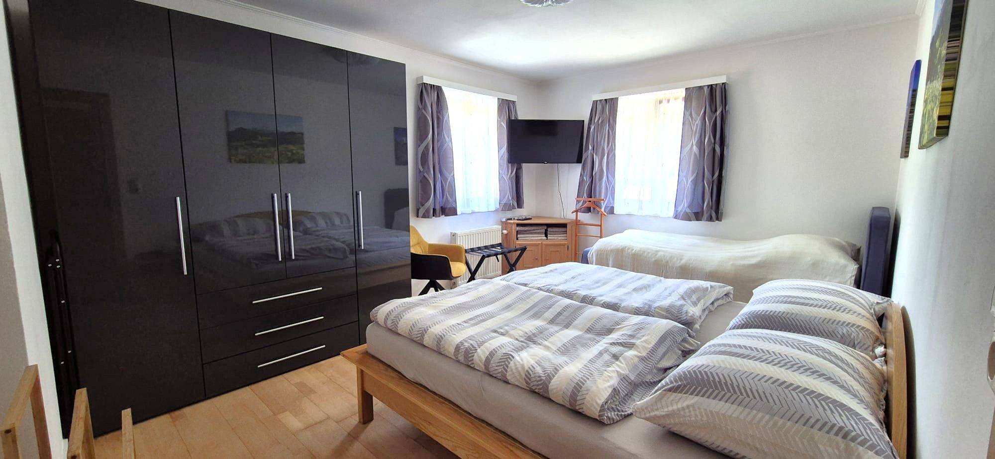 Schlafzimmer mit zwei Betten, einem großen schwarzen Kleiderschrank, einem Fernseher und zwei Fenstern mit Vorhängen.