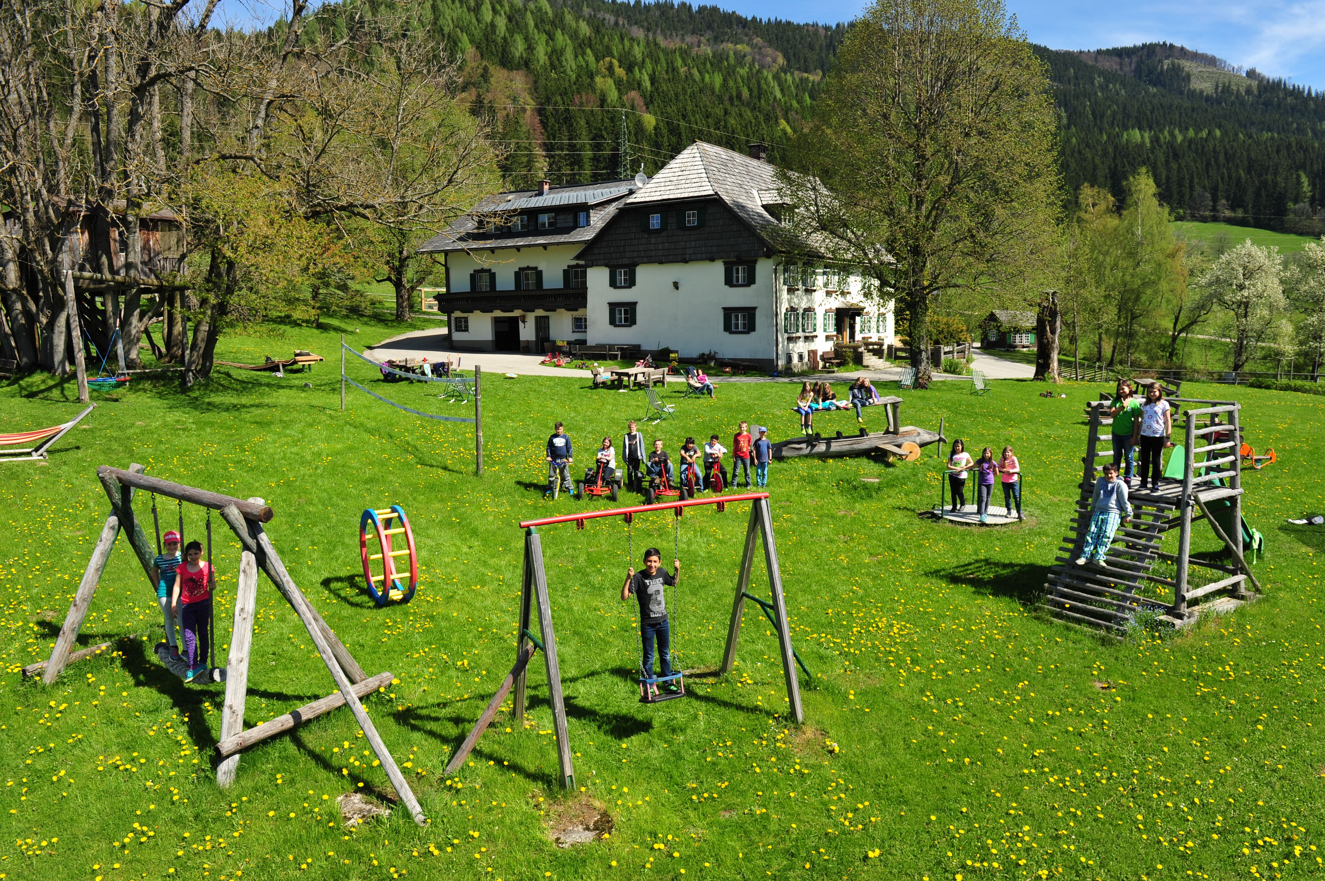 Kinder spielen auf einem Spielplatz vor einem großen Haus in einer grünen Landschaft.