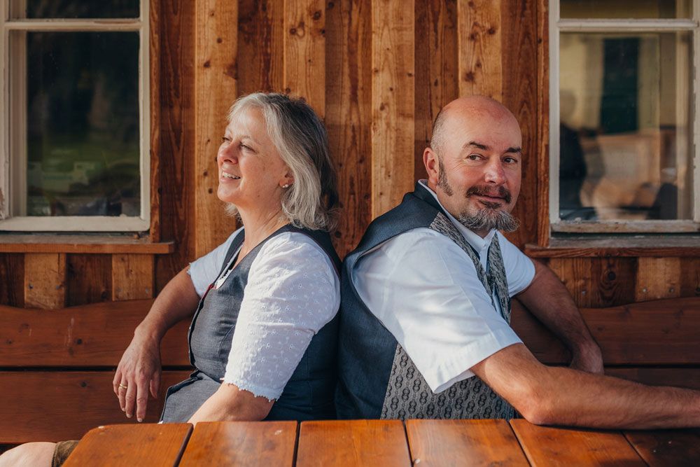 Ein Mann und eine Frau sitzen Rücken an Rücken auf einer Holzbank vor einem Holzgebäude.