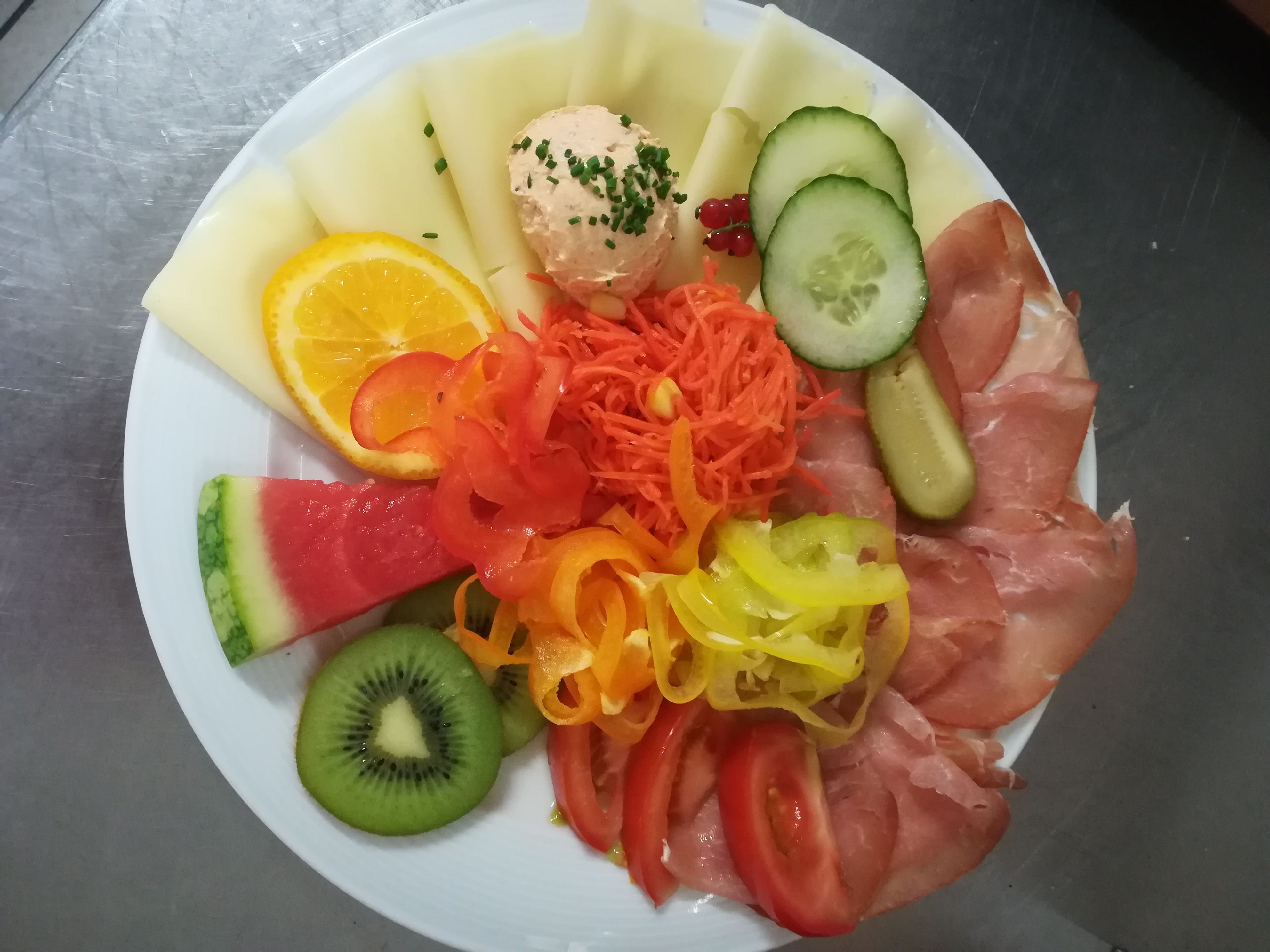Ein Teller mit Käse, Schinken, Gemüse und Obst, darunter Melone, Kiwi, Gurke und Paprika.