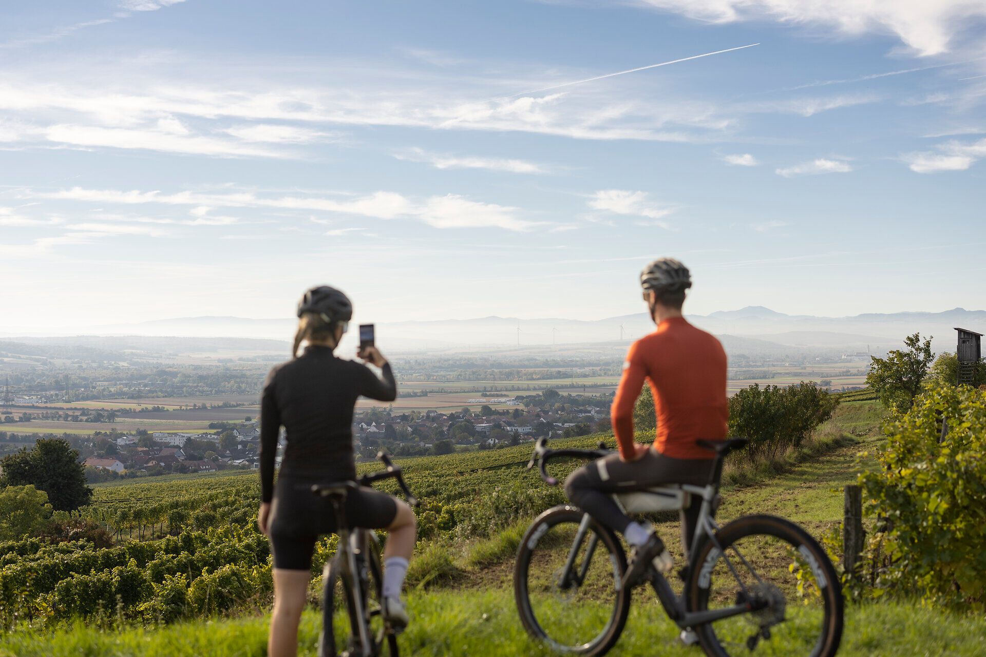 Zwei Radfahrer genießen die atemberaubende Aussicht auf die sanften Hügel und das weite Tal, während die Morgensonne den Himmel in warmen Farben erleuchtet. Die frische Luft und die ruhige Umgebung laden dazu ein, die Schönheit der Natur zu erleben und die Seele baumeln zu lassen.