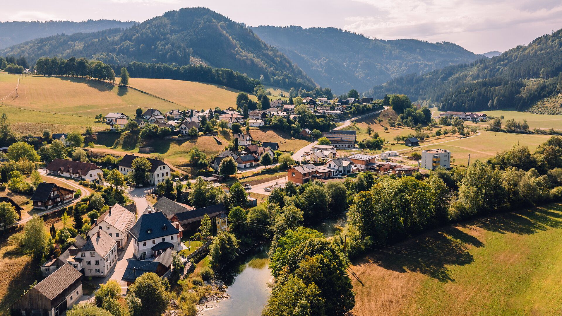 Drohnenaufnahme von Kogelsbach mit dem Ortszentrum, grünen Wiesen und bewaldeten Bergen im Hintergrund.