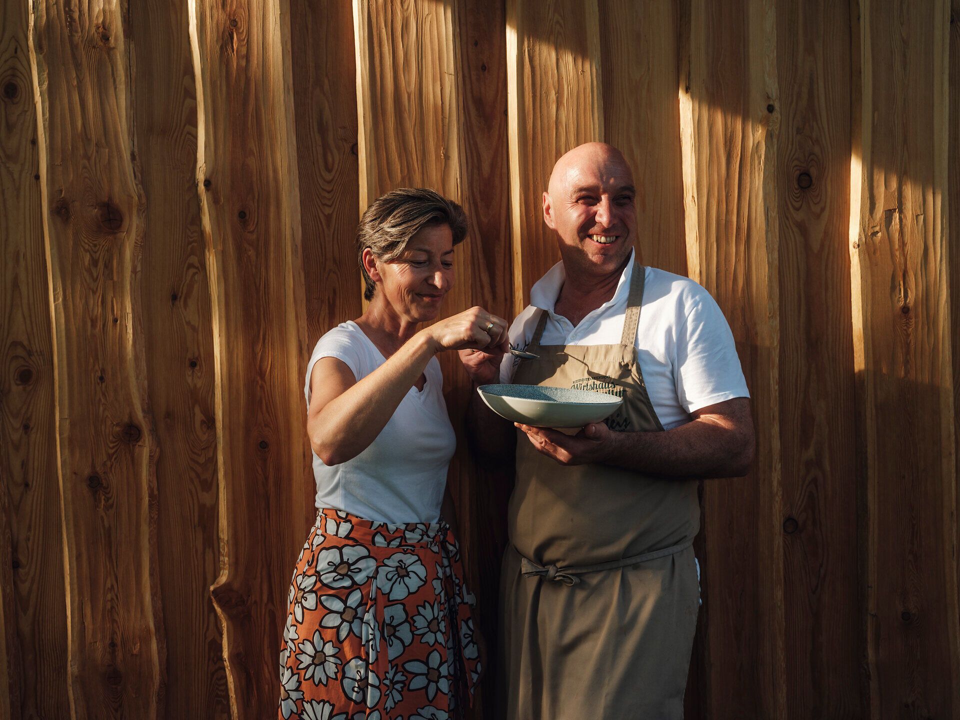 In einer warmen, einladenden Atmosphäre genießen zwei Personen gemeinsam eine kulinarische Kreation. Die Freude am Kochen und die Leidenschaft für regionale Zutaten spiegeln sich in ihren lächelnden Gesichtern wider, während die rustikale Holzverkleidung im Hintergrund eine gemütliche Stimmung schafft.