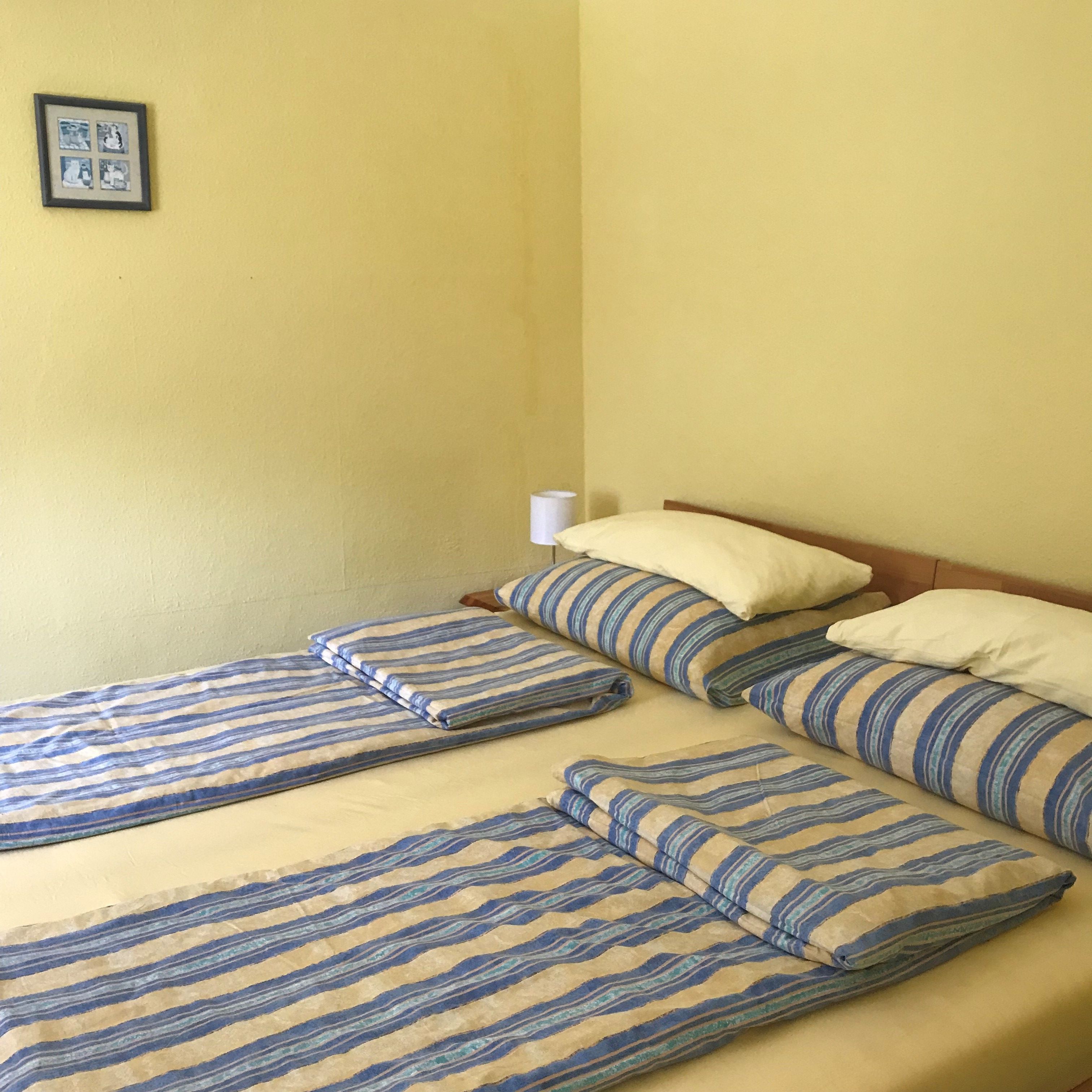Schlafzimmer mit Doppelbett, gestreifte Bettwäsche, gelbe Wände, kleines Bild an der Wand.