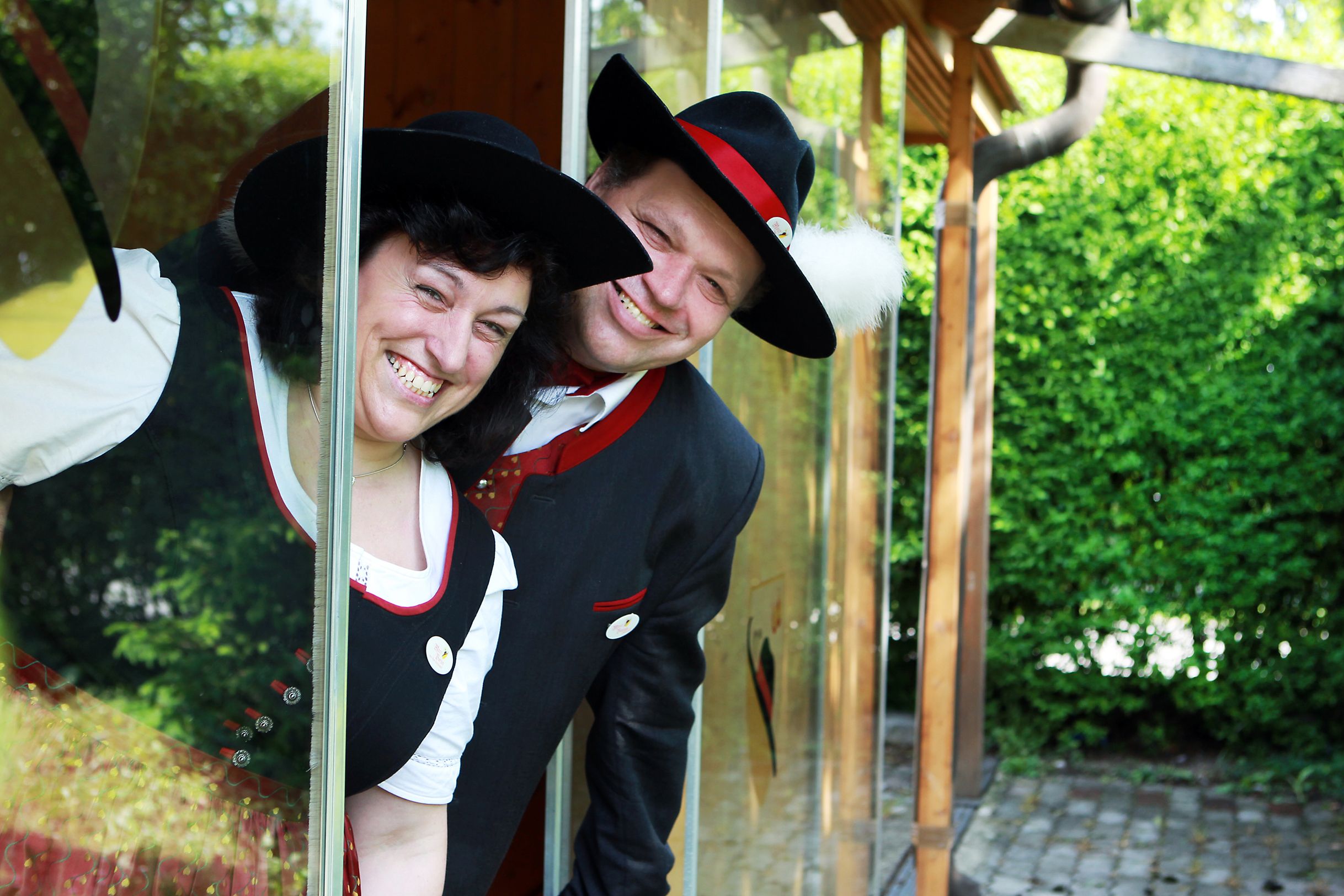 Zwei Personen in traditioneller Tracht lächeln durch ein Fenster.