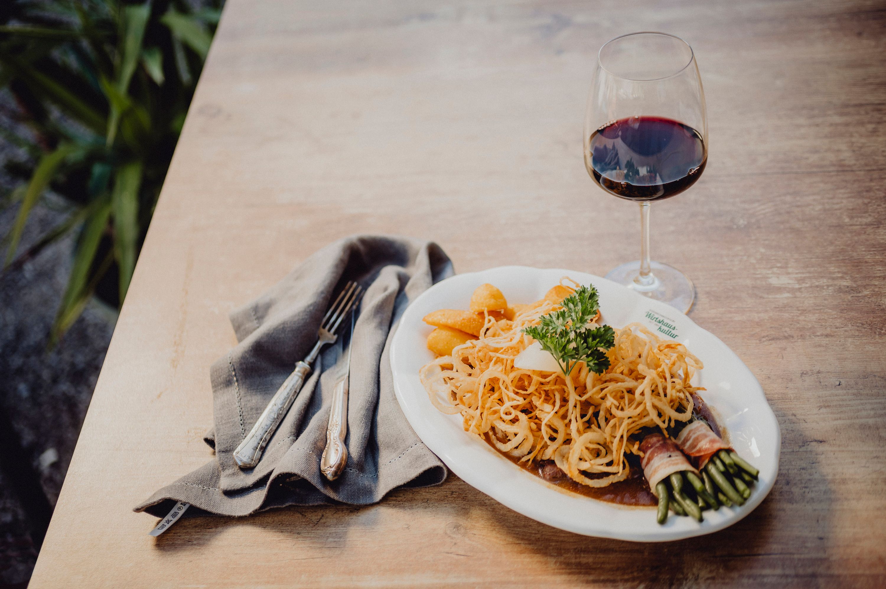 Ein Teller mit Zwiebelrostbraten, Kartoffeln und Speckbohnen, daneben ein Glas Rotwein auf einem Holztisch.
