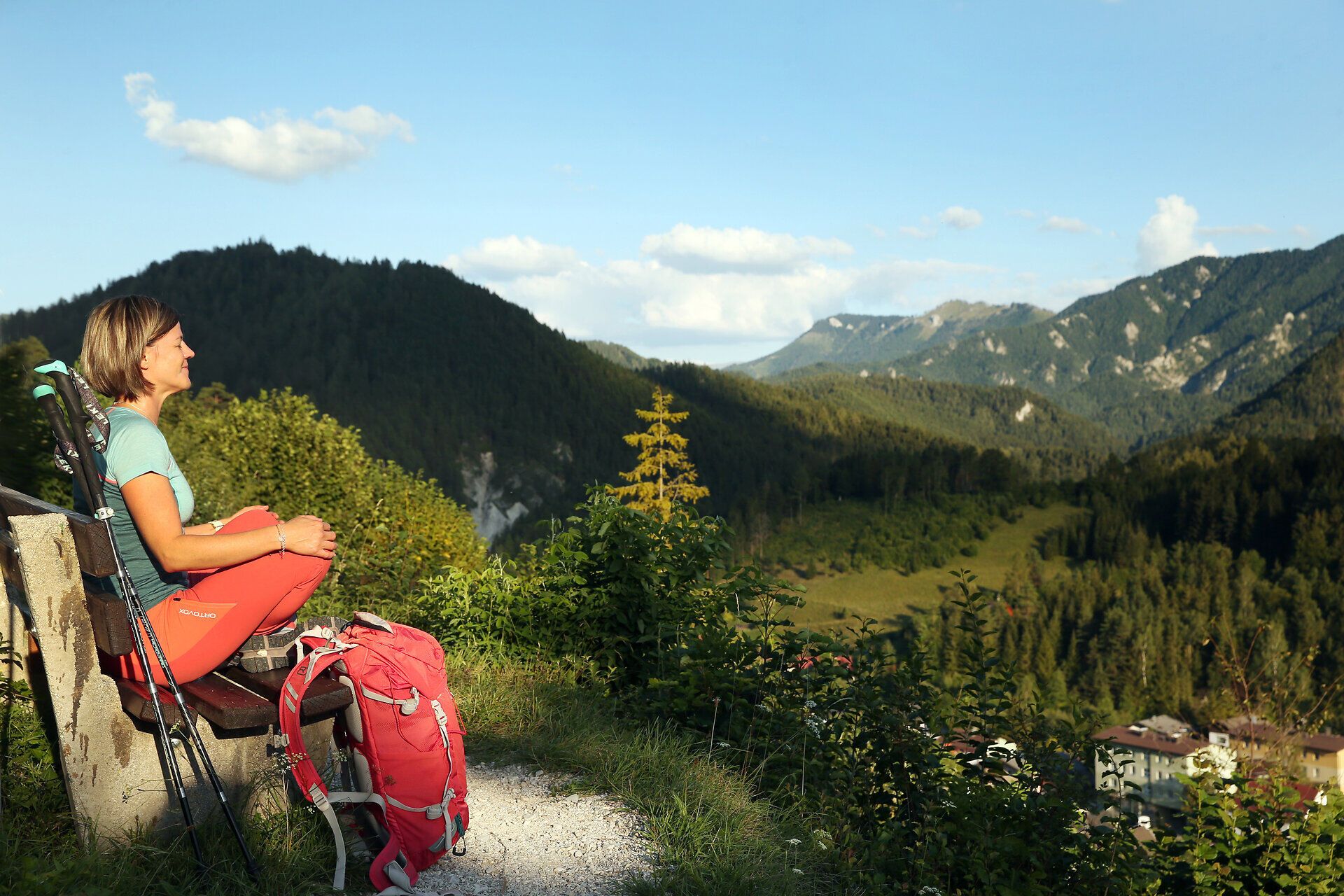 Inmitten der majestätischen Berge sitzt eine Wanderin auf einer Bank und genießt die atemberaubende Aussicht. Die sanften Hügel und dichten Wälder laden zu einer Pilgerwanderung ein, während die frische Bergluft die Sinne belebt. Ein perfekter Moment der Ruhe und Besinnung in der Natur.