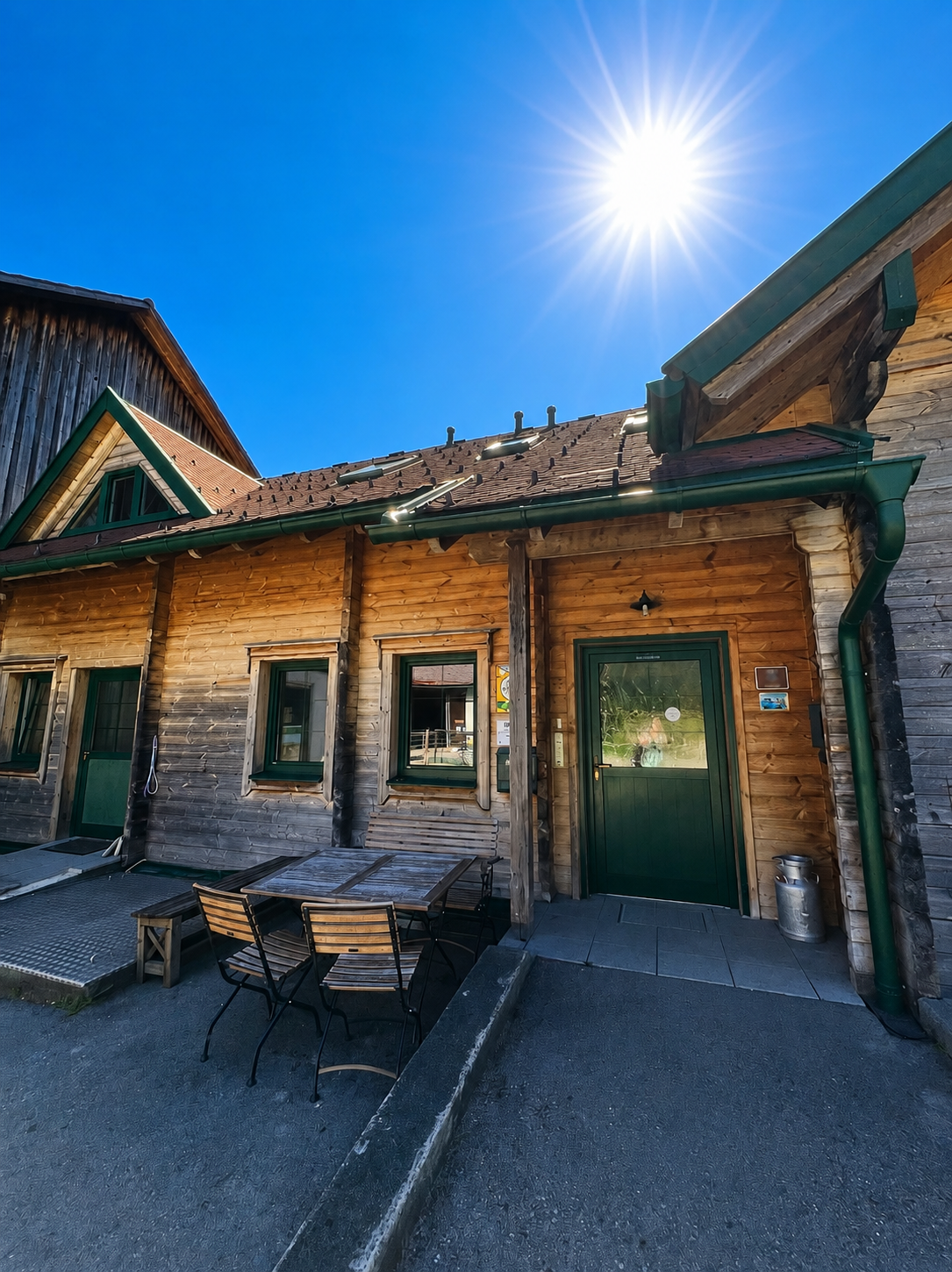 Holzgebäude mit grünem Dach und Tisch mit Stühlen im Vordergrund, unter strahlender Sonne.