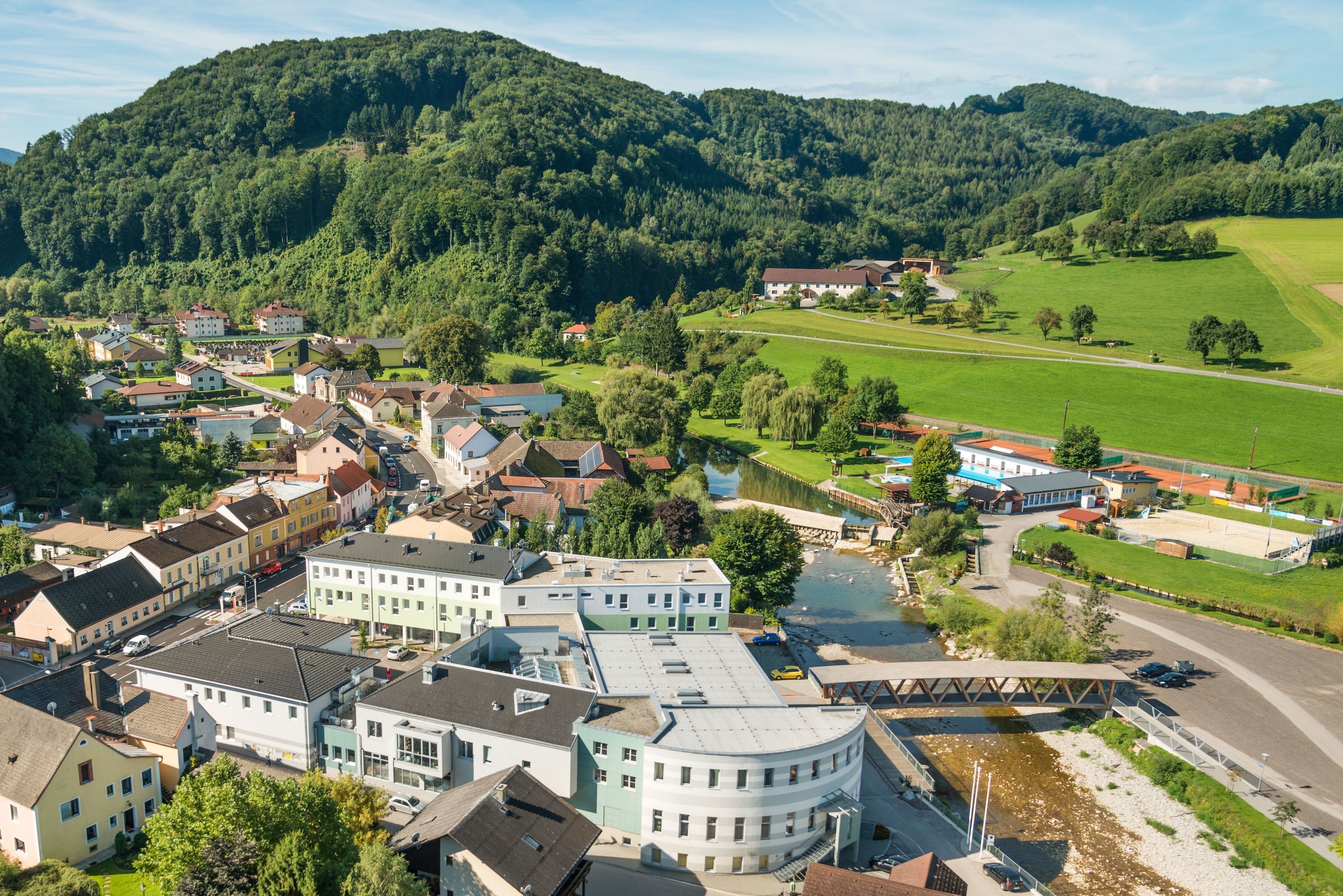 Luftaufnahme eines kleinen Dorfes mit Fluss, Brücke und umliegenden Hügeln.