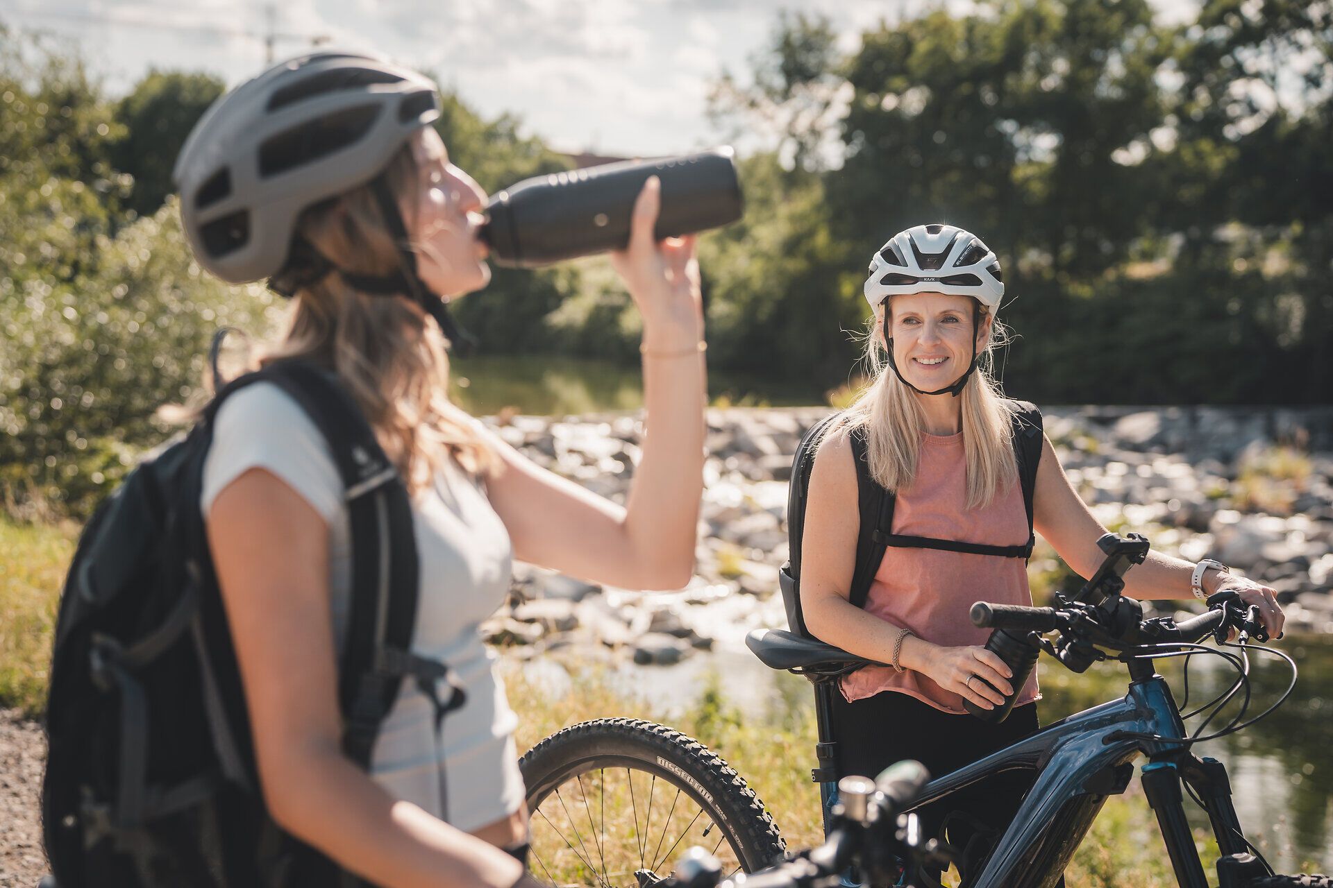 Zwei Radfahrerinnen mit Helmen und Rucksäcken machen mit ihren Rädern Pause an einem Fluss; eine trinkt aus einer Trinkflasche.
