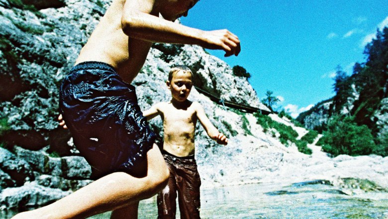 Zwei Kinder spielen im Wasser eines Gebirgsbaches, umgeben von Felsen und blauem Himmel.