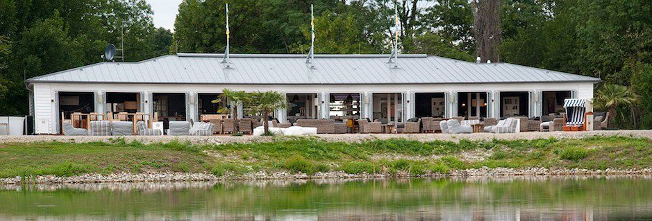 Ein langes Gebäude mit Terrassenmöbeln vor einem Teich, umgeben von Bäumen.
