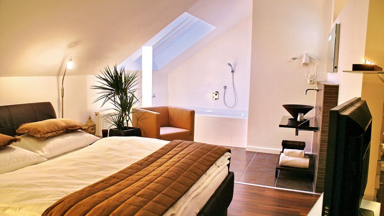 Gemütliches Schlafzimmer mit Doppelbett, Pflanzen, Sessel und Badewanne unter Dachschräge.