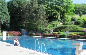 Woman sitting at the edge of an outdoor swimming pool with forest in the background.
