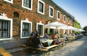 Menschen sitzen an Tischen vor einem Ziegelgeb&auml;ude mit Sonnenschirmen.