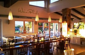 Ein gem&uuml;tliches Gastzimmer mit Holztischen und St&uuml;hlen, dekoriert mit Kerzen und Pflanzen, beleuchtet von warmem Sonnenlicht.