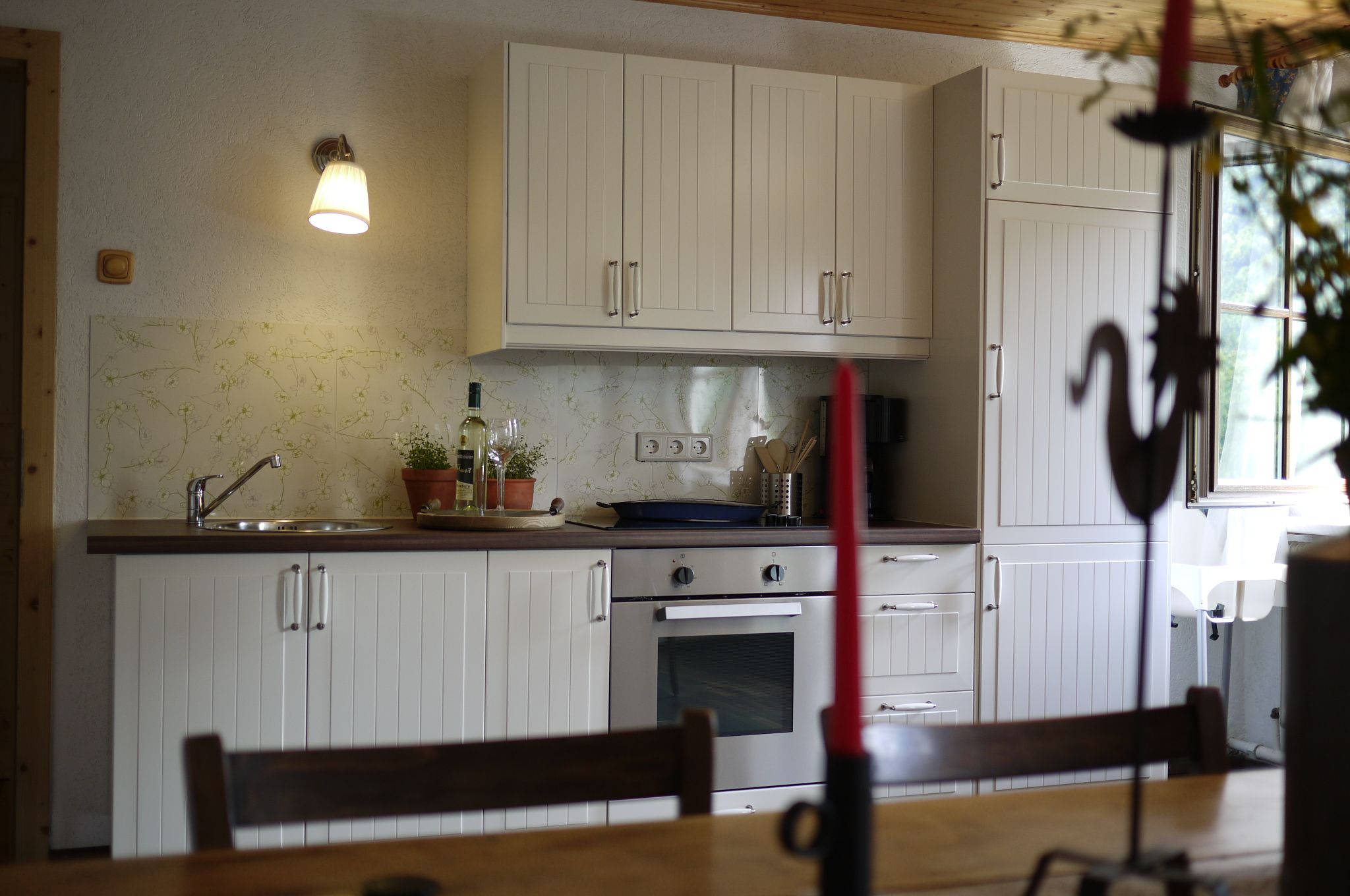 Küche mit weißen Schränken, Herd und Spüle, dekoriert mit Pflanzen und einer Weinflasche.