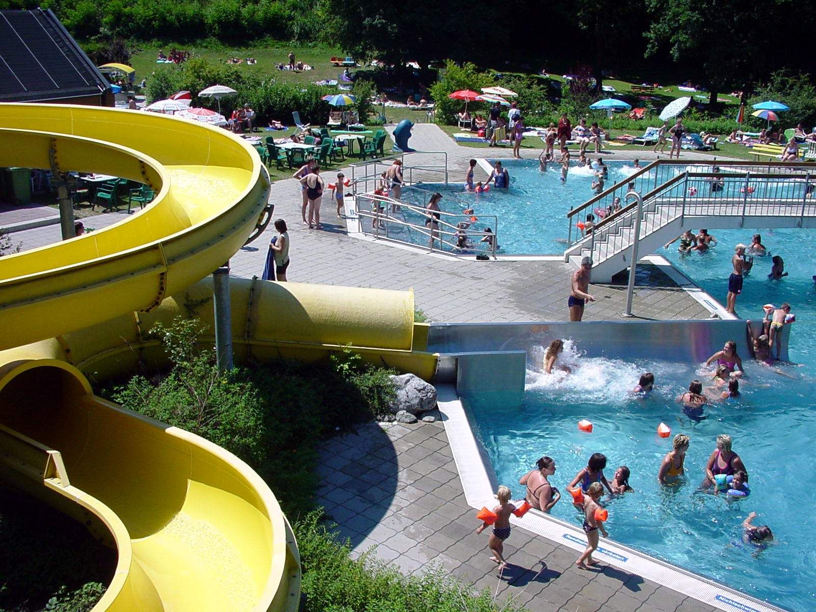 Ein Freibad mit gelber Wasserrutsche, Schwimmern im Becken und Menschen auf der Liegewiese.