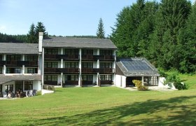 Hotelgebäude mit Solarpanelen und grüner Wiese im Vordergrund, umgeben von Wald.