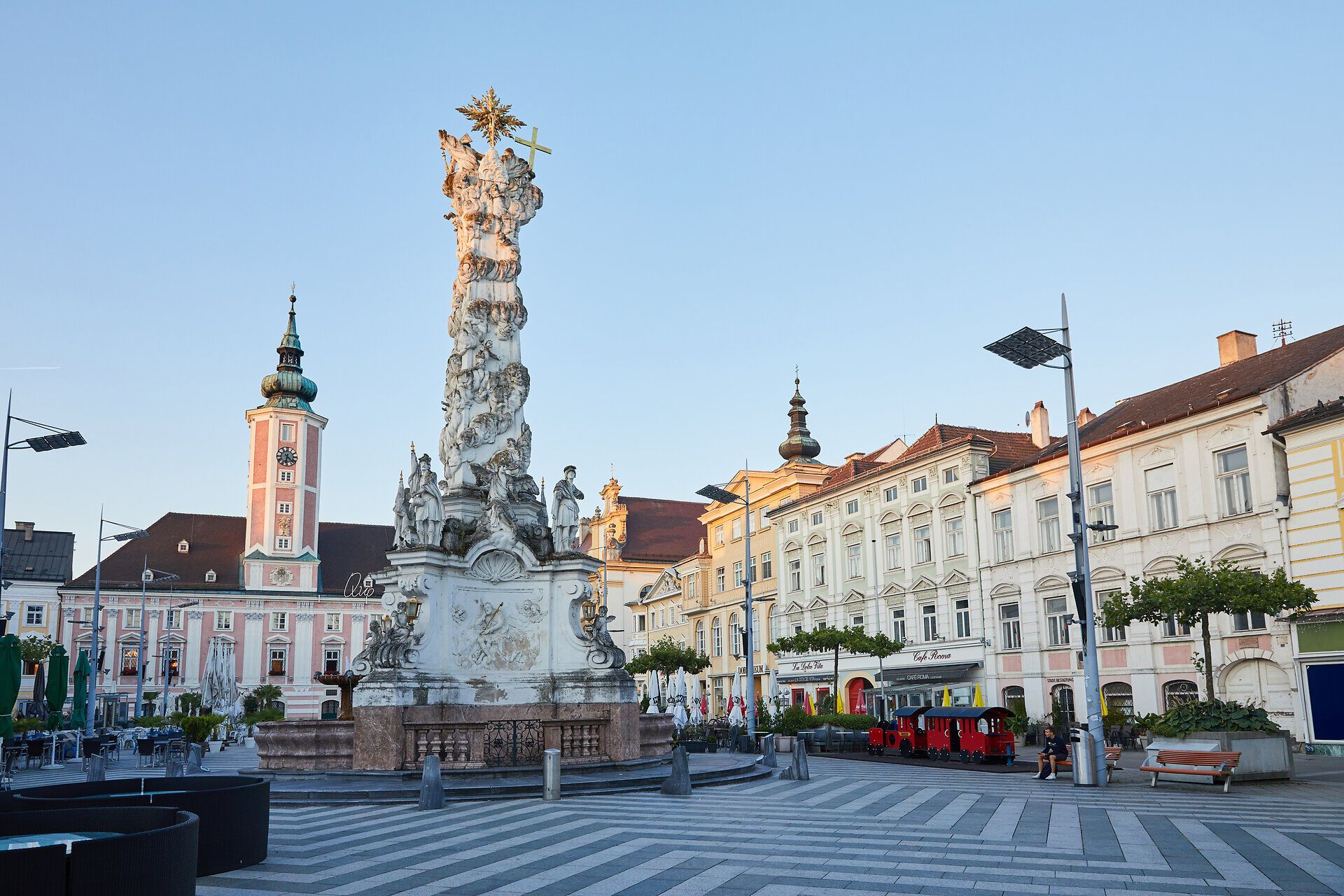 Der Platz strahlt eine ruhige Eleganz aus, umgeben von historischen Gebäuden und dem beeindruckenden Denkmal in der Mitte. Die sanften Morgenstrahlen tauchen die Szene in ein warmes Licht und laden dazu ein, die Atmosphäre der Stadt zu genießen.