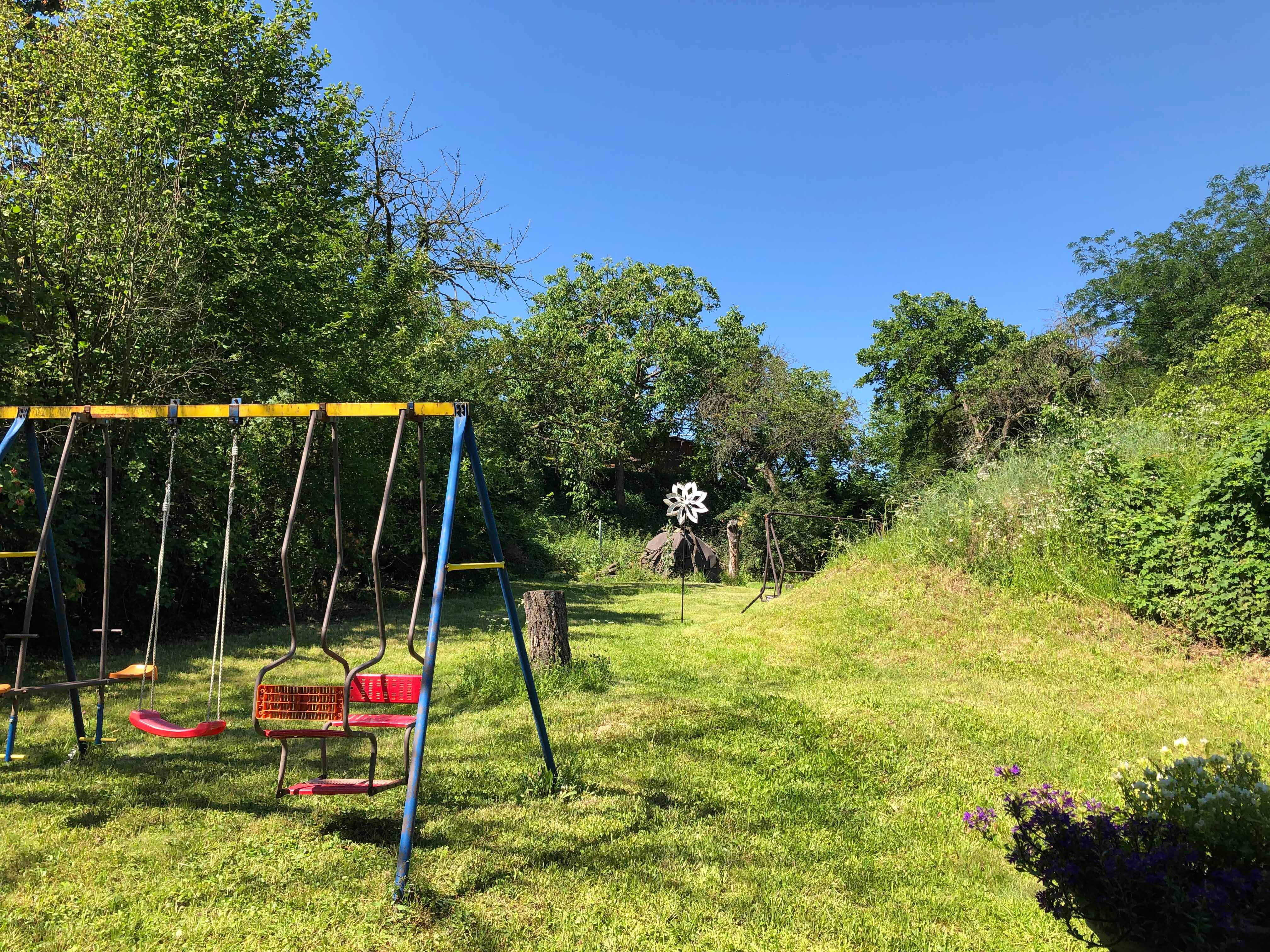 Ein Garten mit einer Schaukel, umgeben von Bäumen und Wiese, unter blauem Himmel.