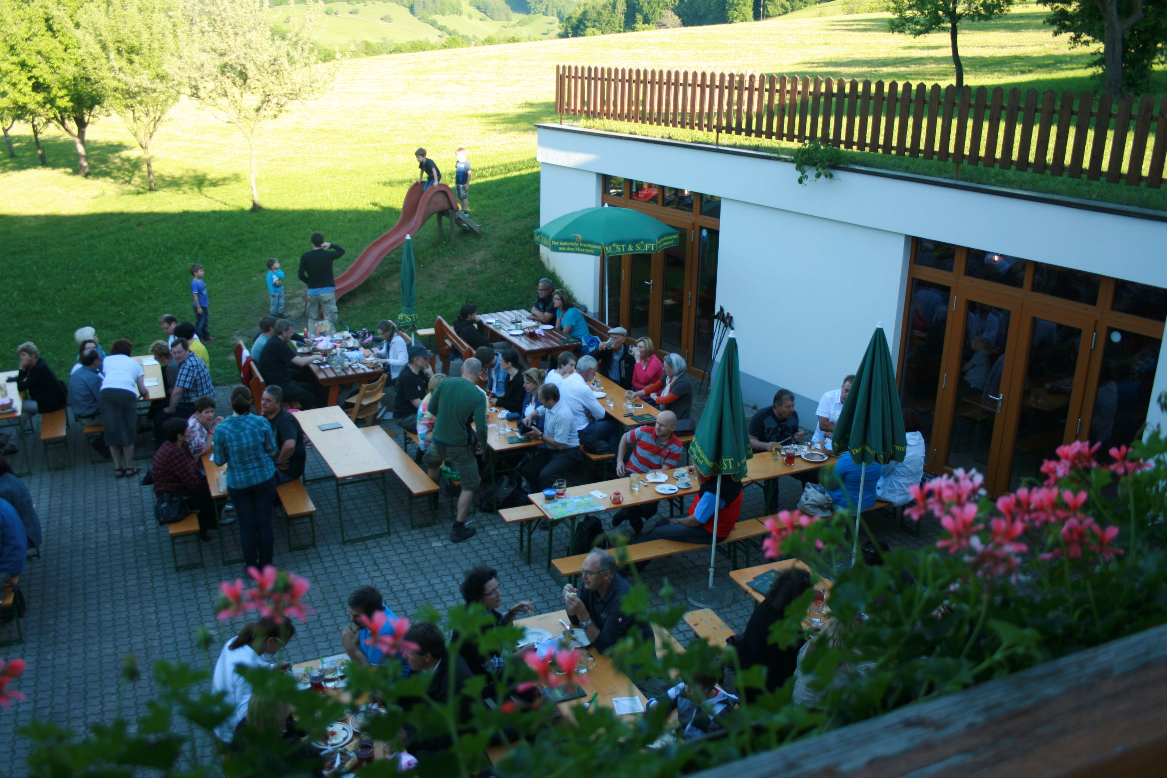 Menschen sitzen an Tischen im Freien, umgeben von grüner Landschaft und Blumen.