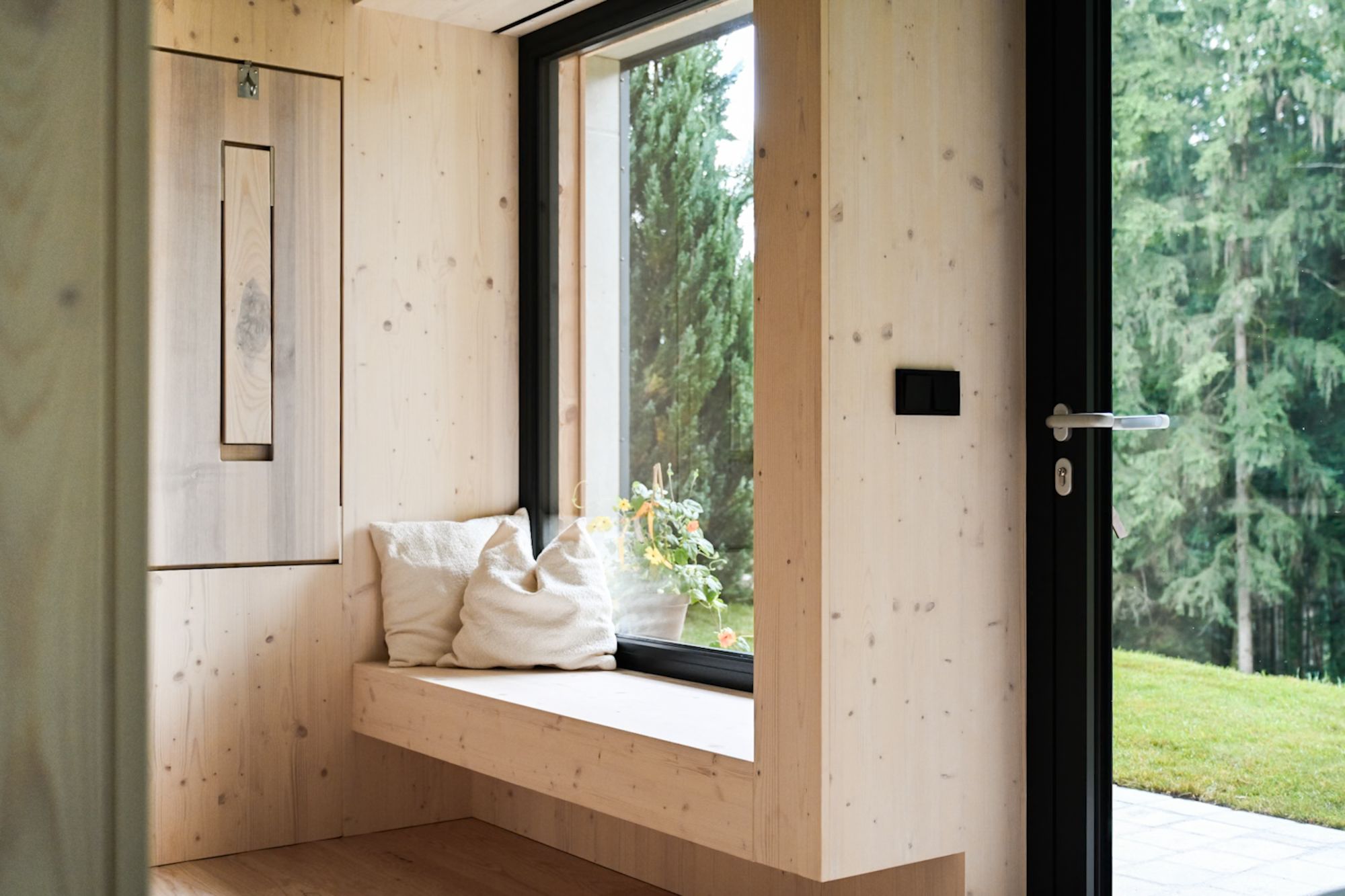 Innenansicht eines modernen Holzhauses mit großem Fenster und Sitzbank, Blick auf einen Wald.