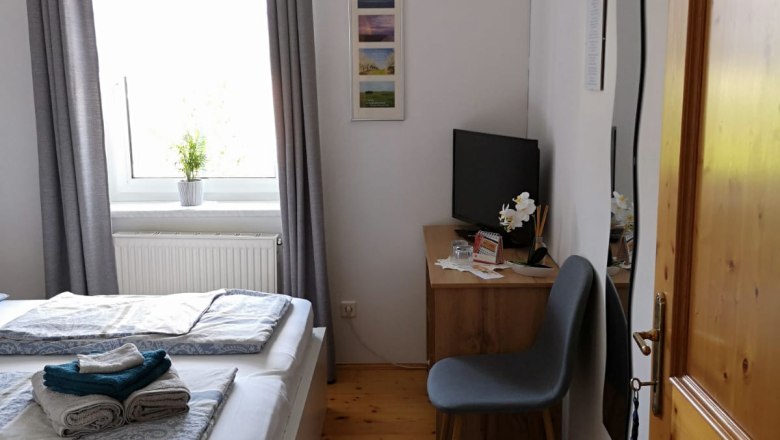Bright bedroom with double bed, gray curtains, small desk and chair, TV and carpet on wooden floor.