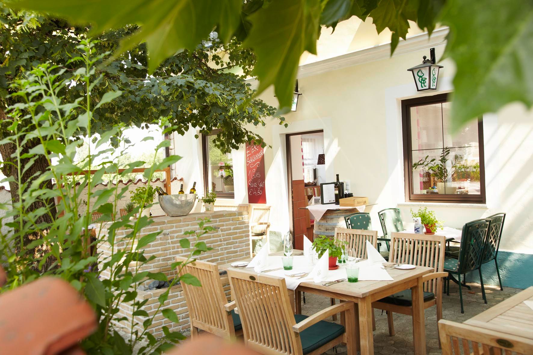 Ein gemütlicher Gastgarten mit Holztischen und Stühlen, umgeben von Pflanzen und Bäumen.