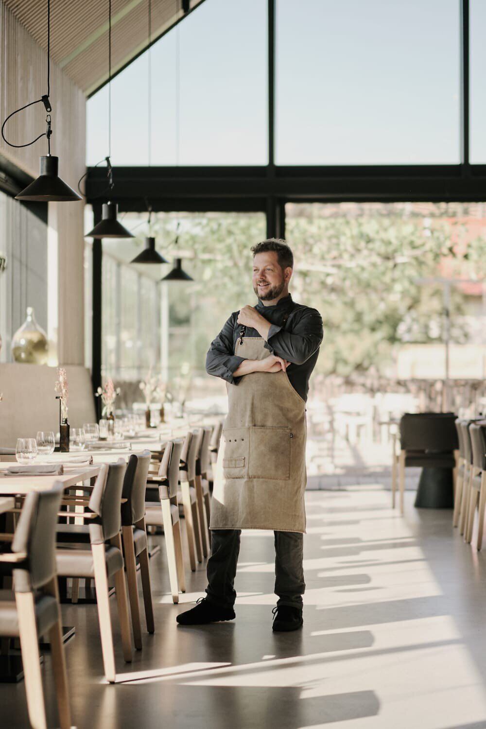In einem lichtdurchfluteten Raum, umgeben von warmen Holztönen und einer einladenden Atmosphäre, steht ein Koch in einem rustikalen Outfit. Die Tische sind liebevoll gedeckt und laden dazu ein, die kulinarischen Köstlichkeiten der Region zu genießen. Hier wird die Leidenschaft für die lokale Küche spürbar.