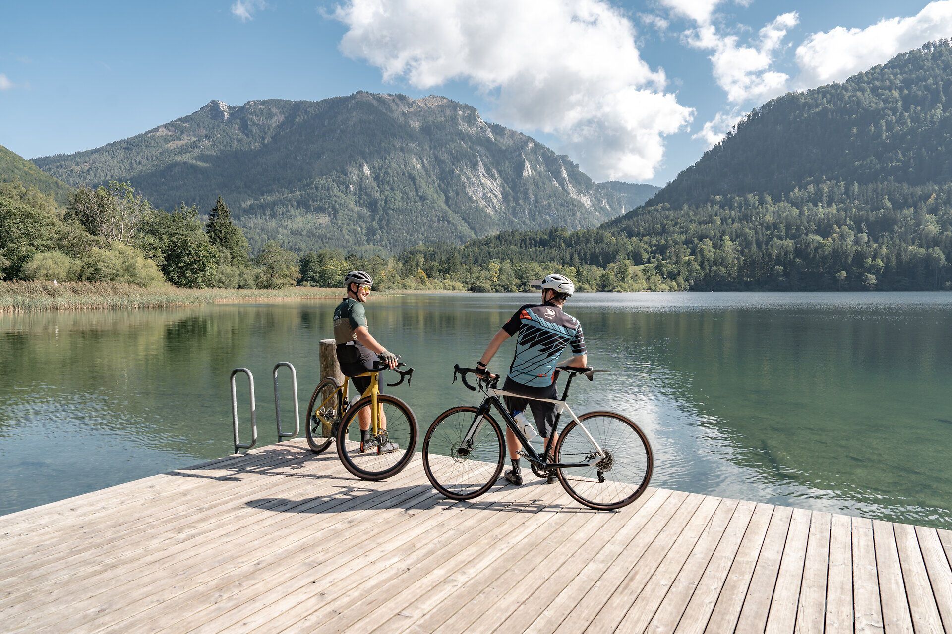 Die sanften Hügel und majestätischen Berge umrahmen den glitzernden See, während zwei Radfahrer auf dem Holzsteg innehalten und die atemberaubende Aussicht genießen. Die frische Luft und die ruhige Atmosphäre laden dazu ein, die Schönheit der Natur zu erkunden und unvergessliche Momente zu erleben.