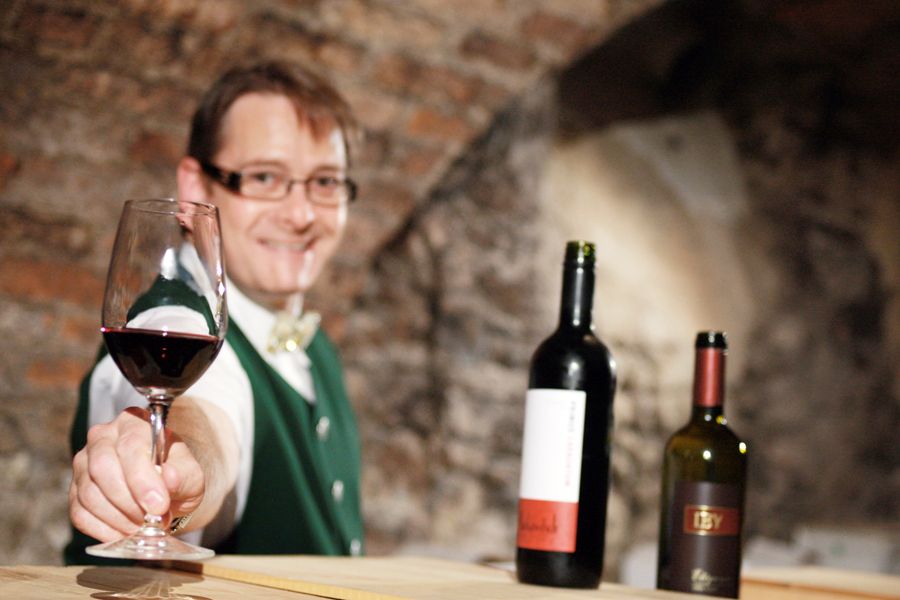 Ein Mann in traditioneller Kleidung bietet ein Glas Rotwein an, im Hintergrund zwei Weinflaschen.